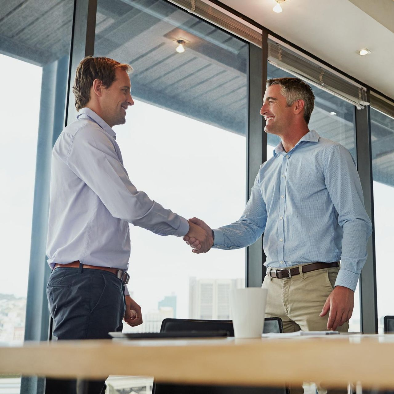Two men in shirts shaking hands in an office, smiling.