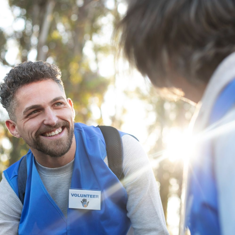 Man smiling, wearing a blue volunteer vest, talking to another person outdoors. Sunlight shines.