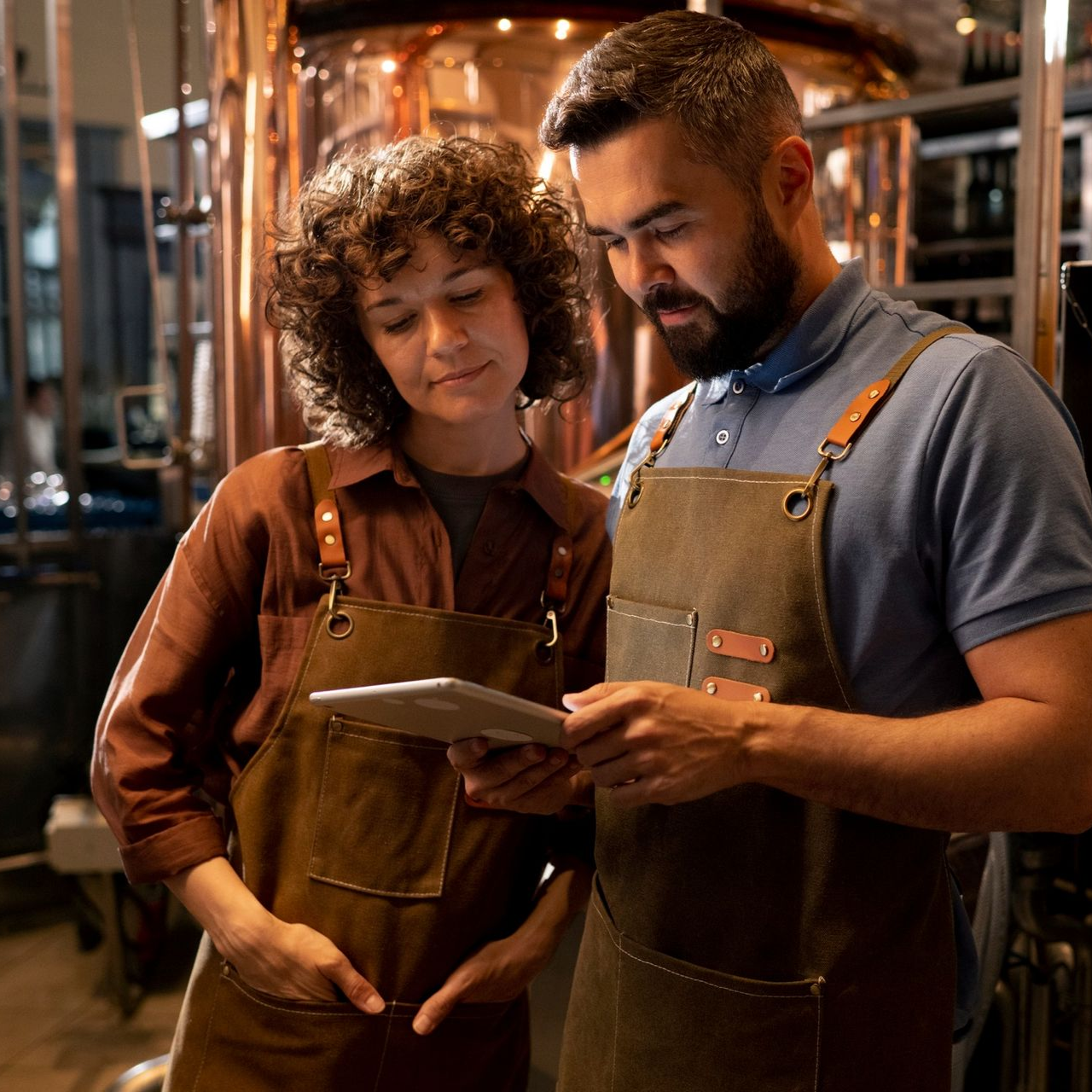 Two people in aprons look at a tablet near copper brewing equipment.