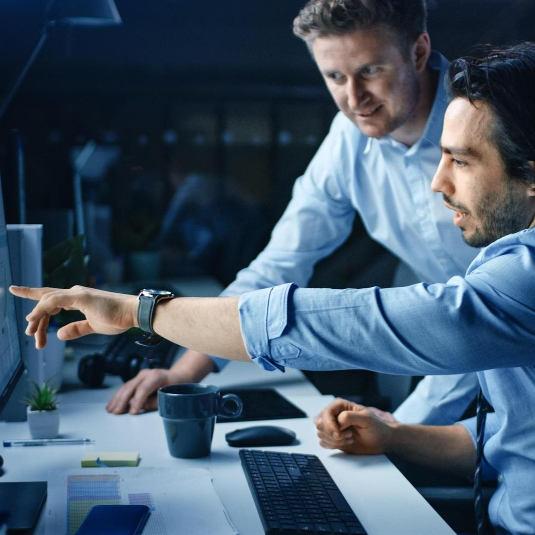 Two men in blue shirts looking at computer screen, one points.