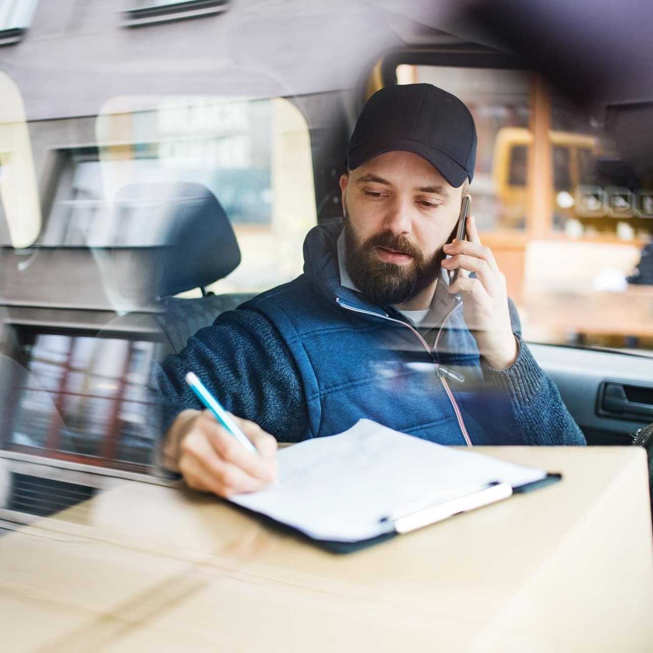 Delivery driver in van on phone, writing on clipboard, next to packages.