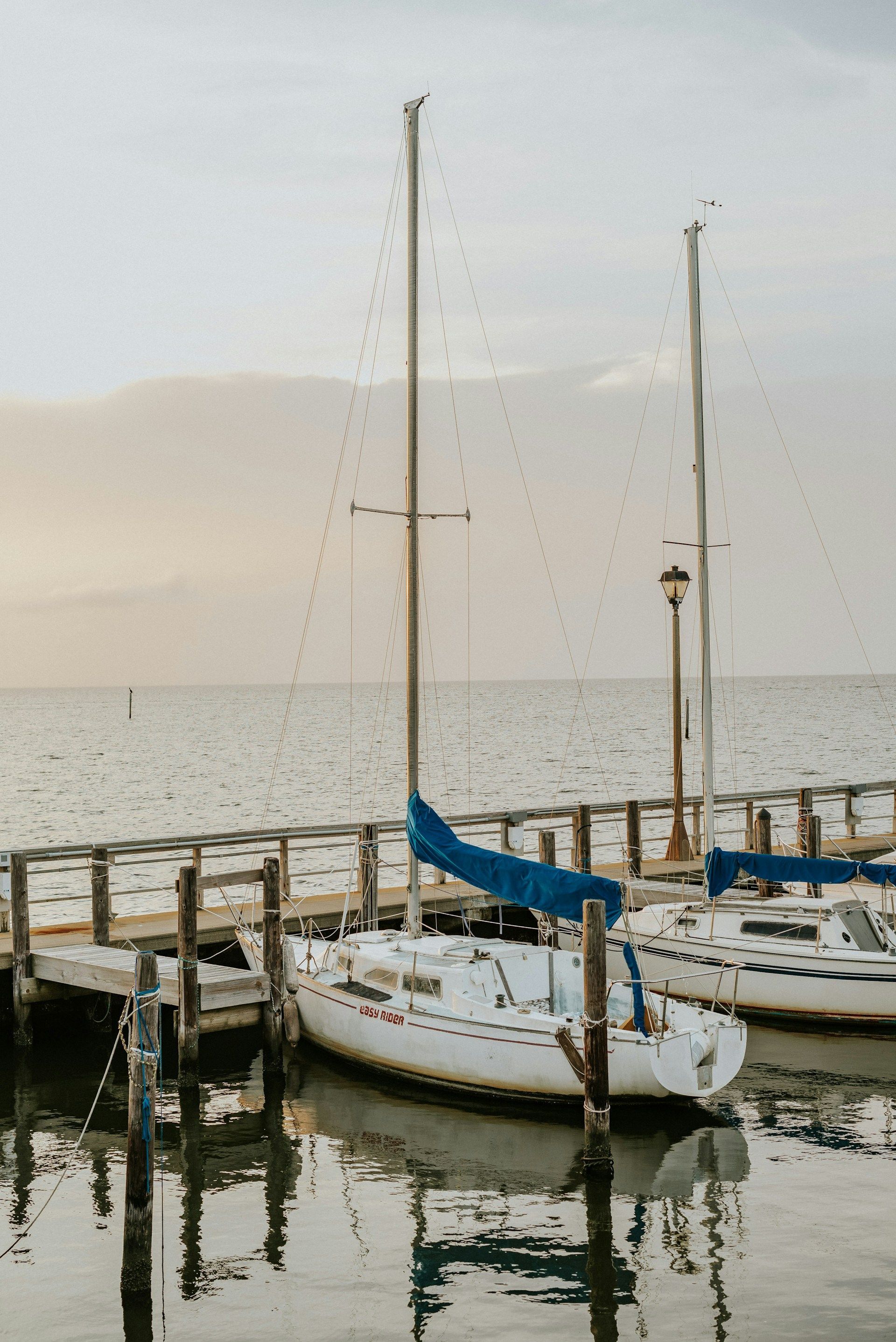Sailboats docked on a wooden pier, reflecting in the water; soft sunlight on the water and sky.