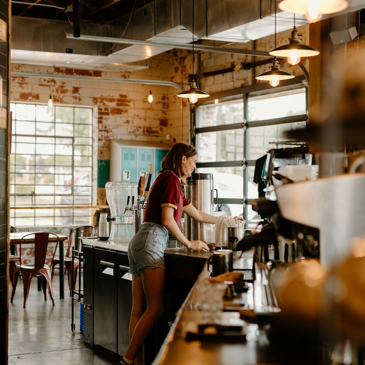 Woman working behind the counter of a coffee shop, with brick walls and large windows.