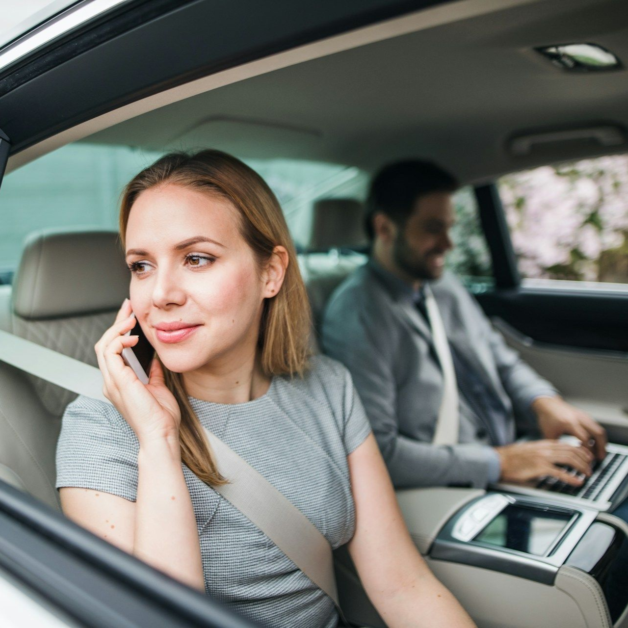 Woman in car talking on phone; man in back seat works on laptop.