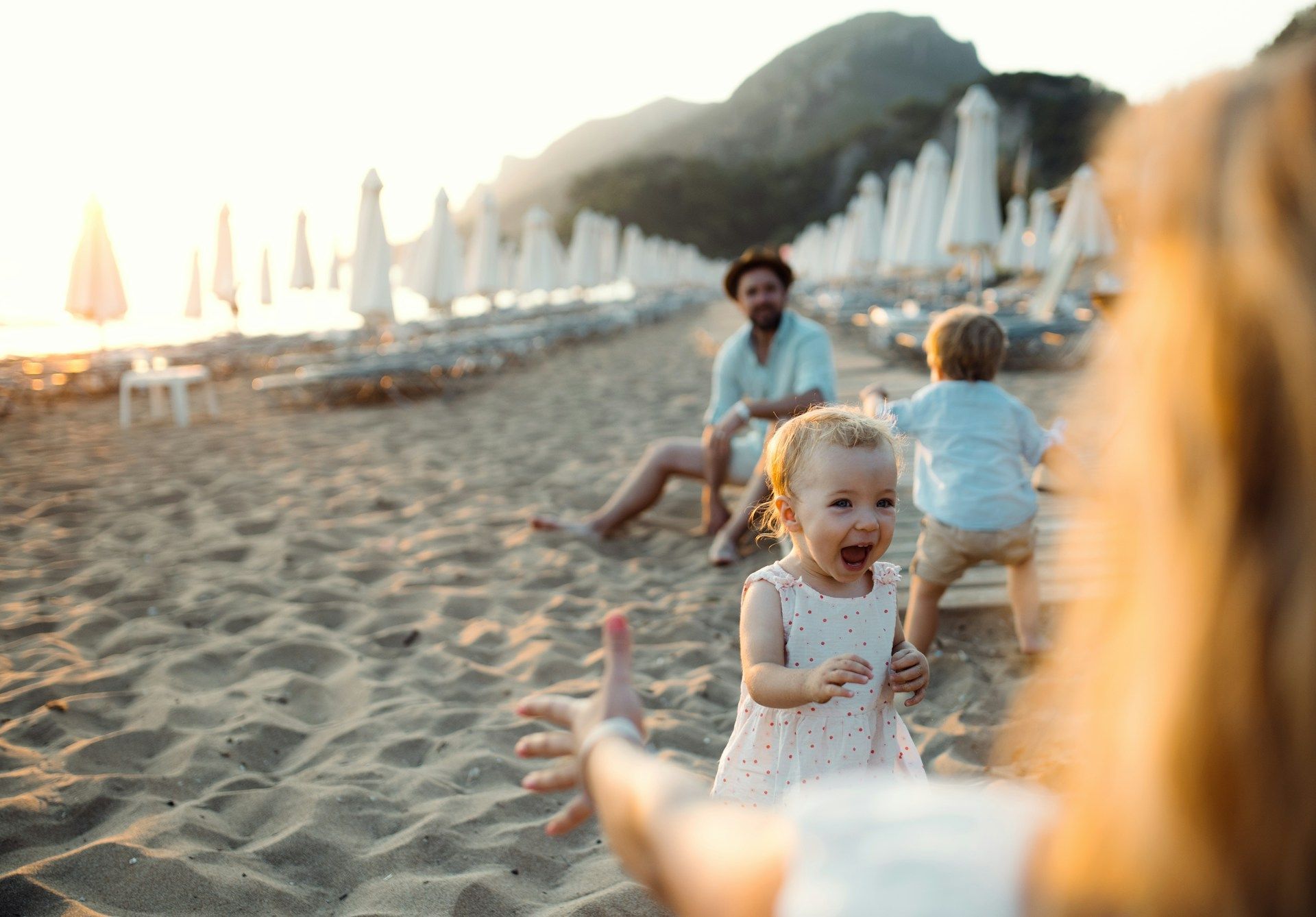 Family playing on a beach. A child runs towards the camera, another child and parent in the background. Sand and umbrellas.