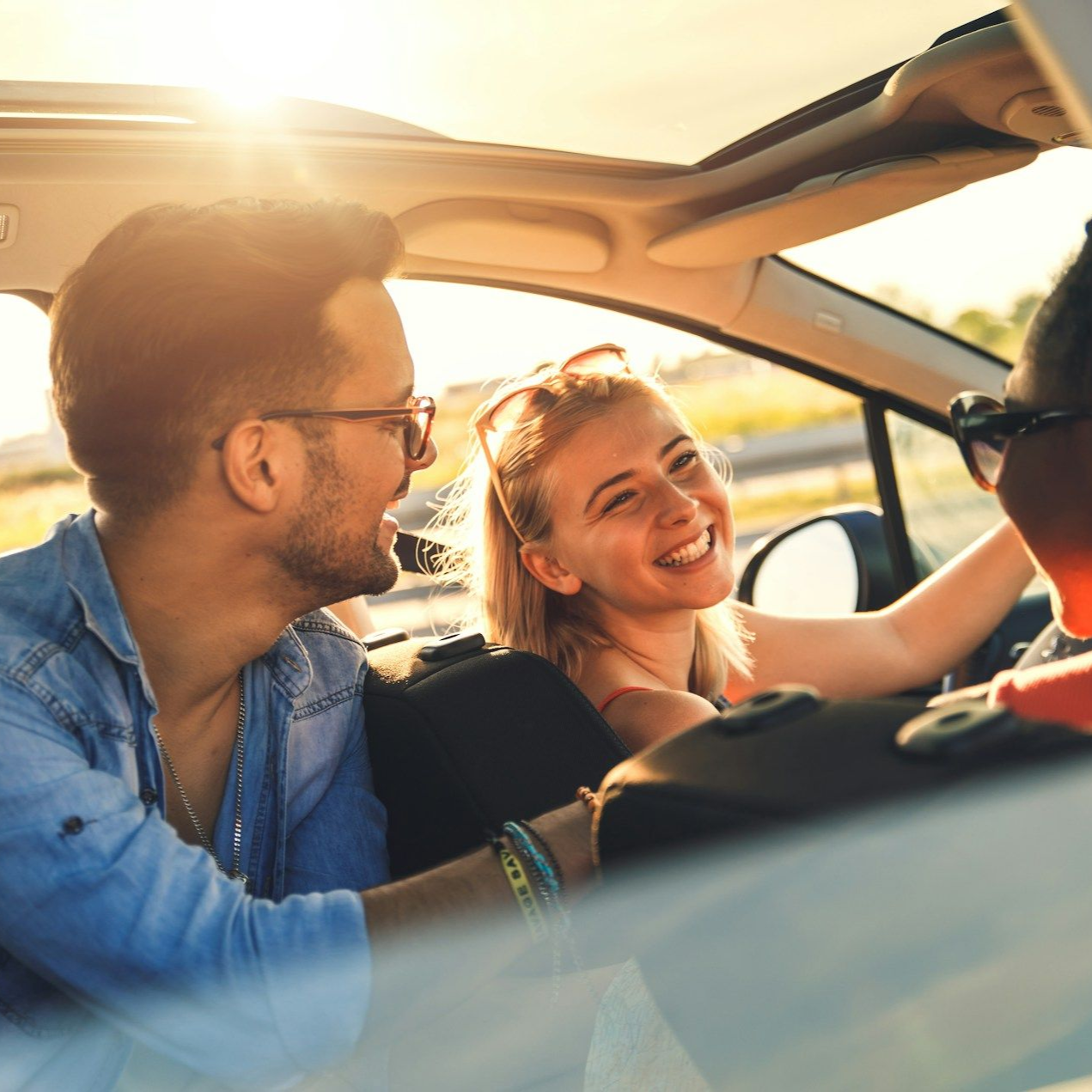 Three people in a car, sunlit, smiling. One driving, the other two in passenger seats.
