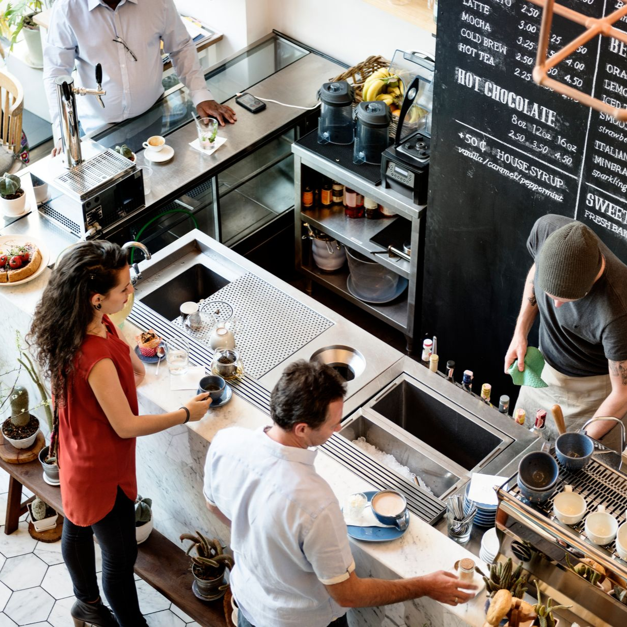 People at a coffee shop counter: one paying, another receiving a drink, two baristas working.