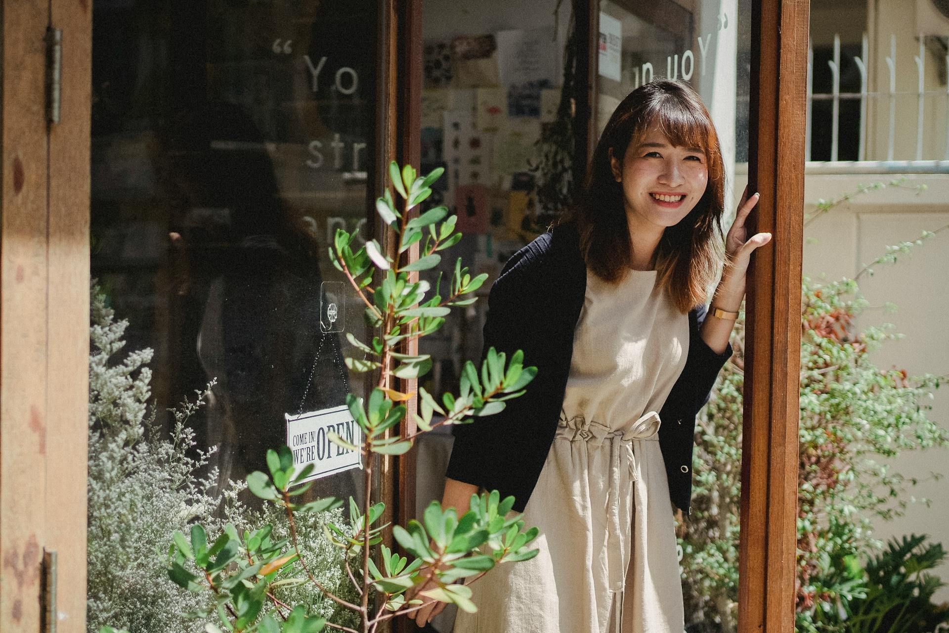 Woman smiling, peeking out of a shop doorway, surrounded by plants; wearing beige dress and black cardigan.