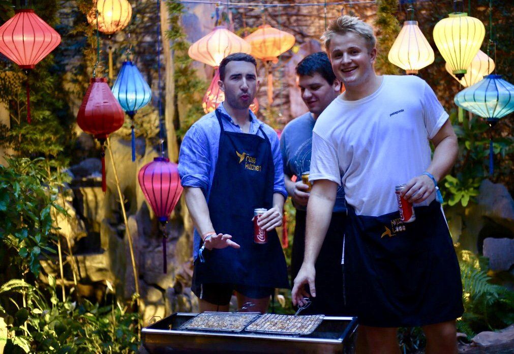 Three men are standing around a grill with lanterns in the background.