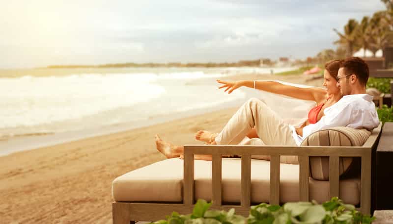 A man and a woman are sitting on a couch on the beach looking at the ocean.