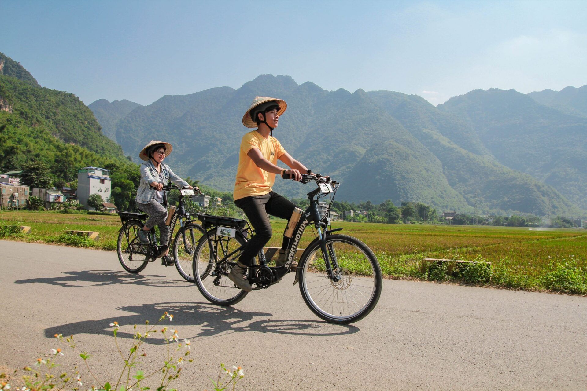 Two people are riding bicycles down a road with mountains in the background.