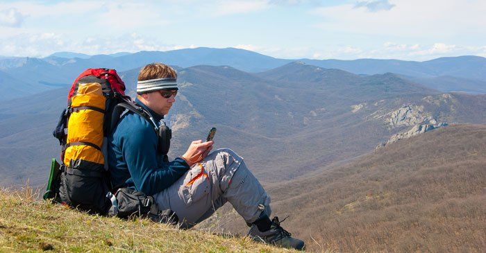A man with a backpack is sitting on top of a mountain looking at his cell phone.