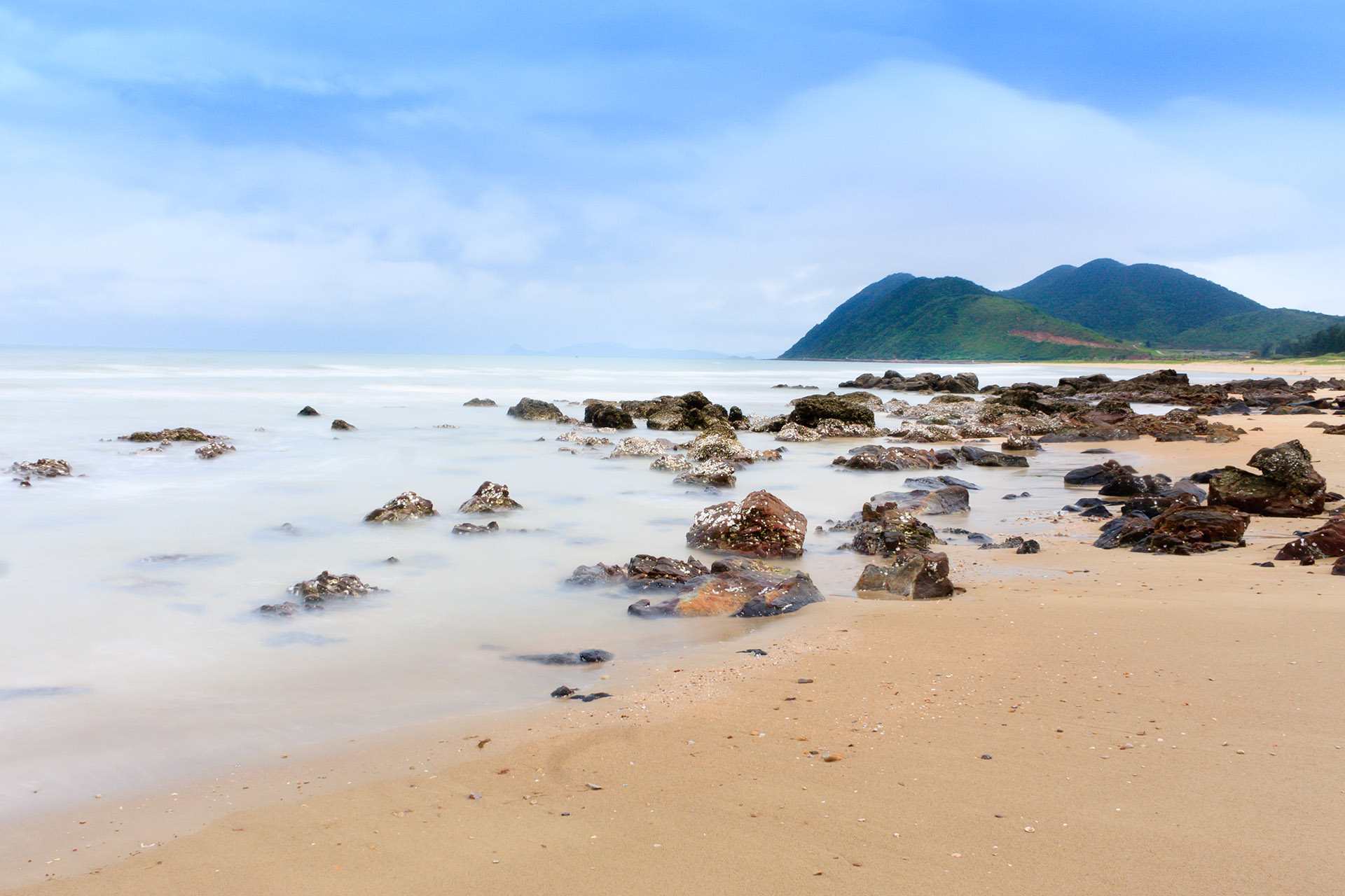 A beach with rocks in the water and a mountain in the background.