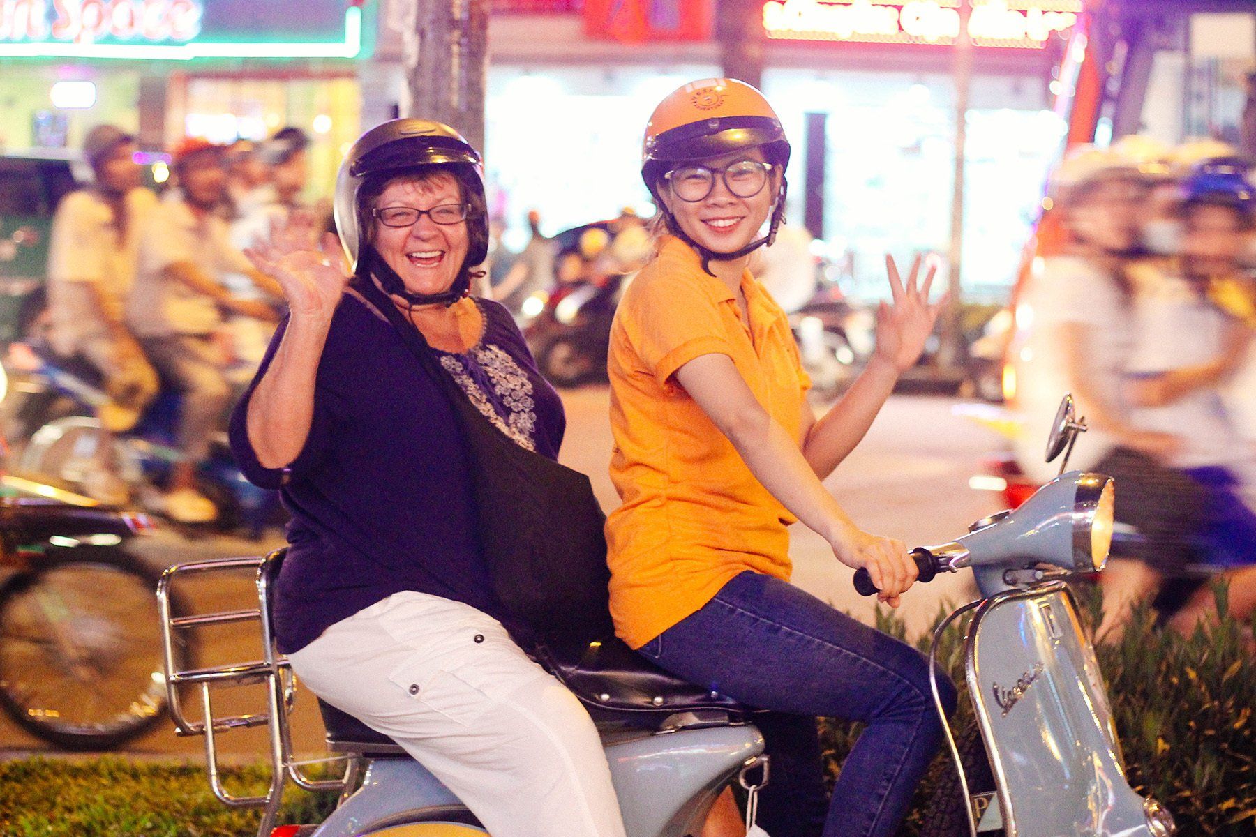 Two women are riding a scooter and waving at the camera.