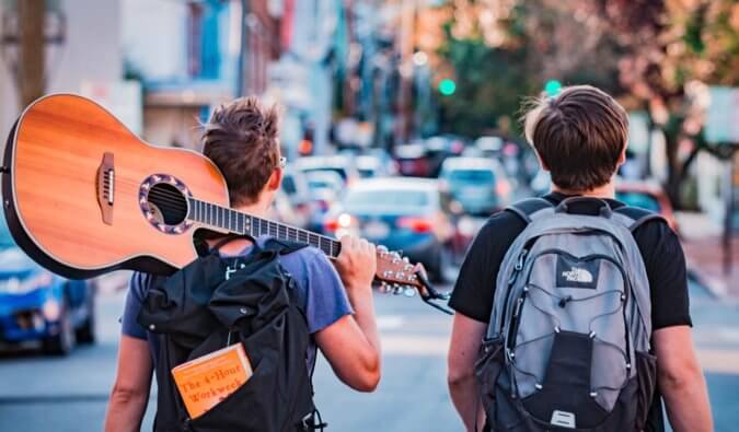Two men with backpacks are walking down a street carrying guitars.