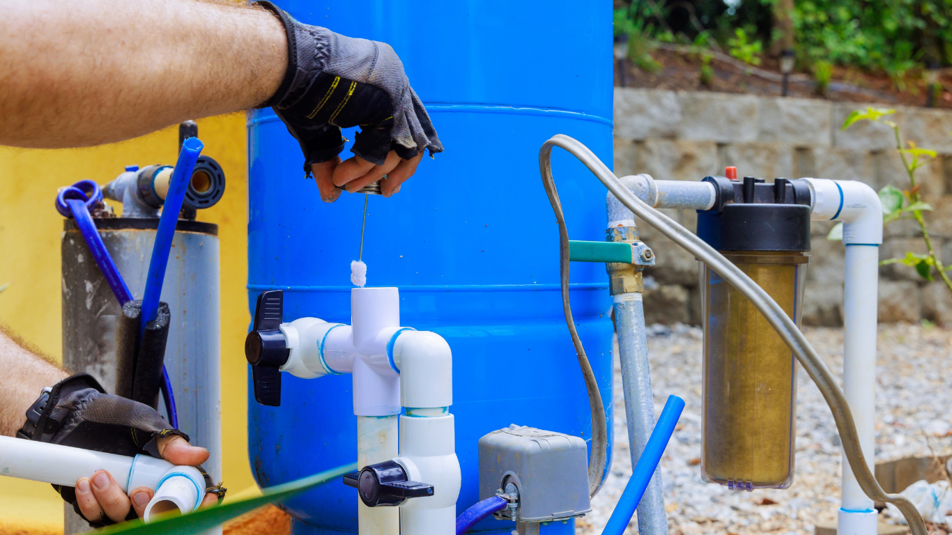 Person in gloves working on a blue water tank with white pipes outside.