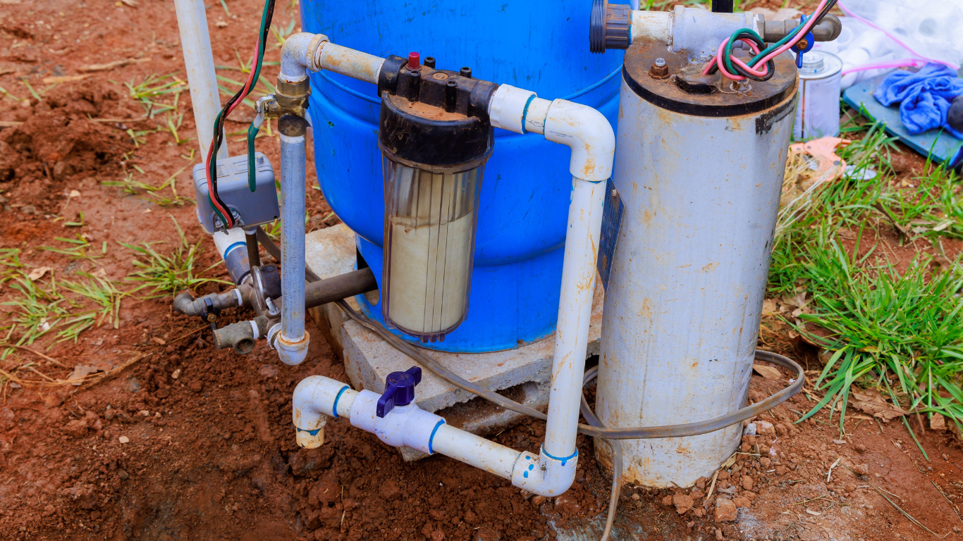 Well water system with blue tank, filter, and plumbing in a dirt setting.