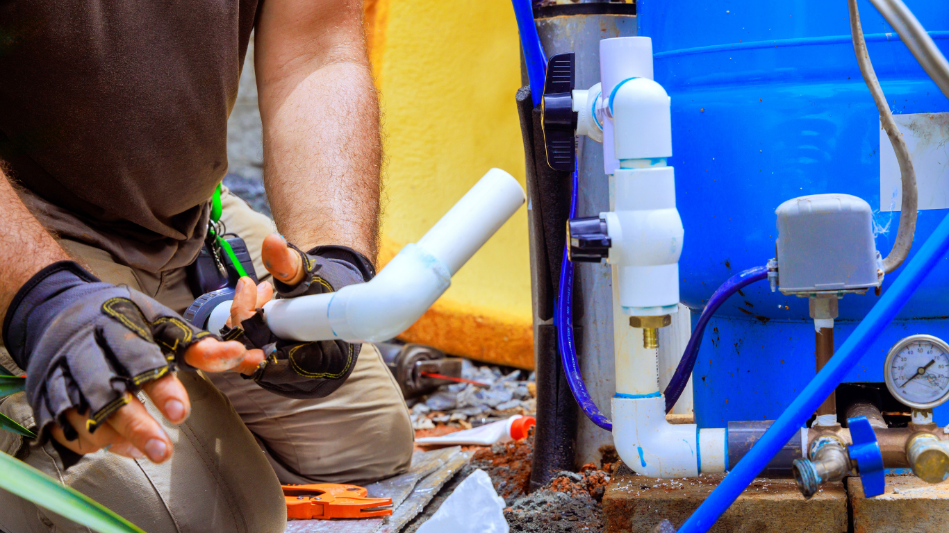 Person in work gloves connecting a white PVC pipe. Blue tank, valves, and gauge are nearby.