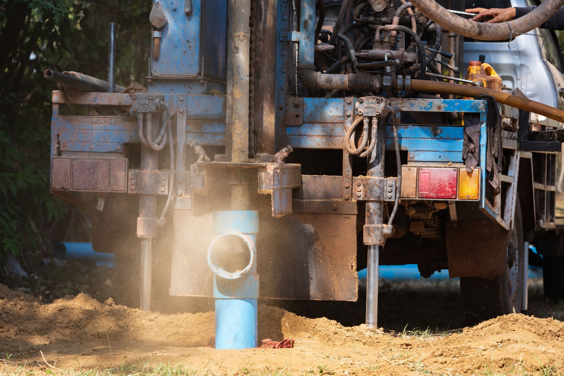 Drilling rig working on a well, emitting dust, with blue pipe and red taillights in a natural setting.
