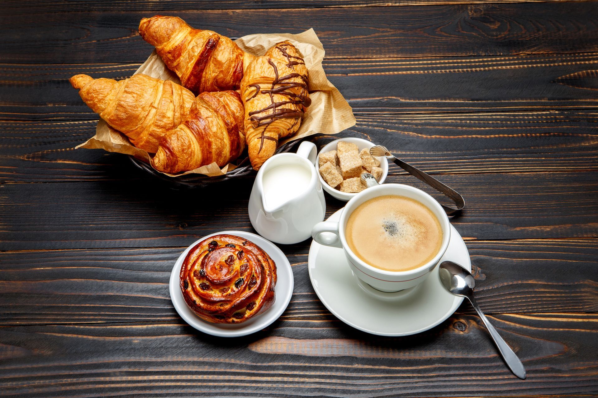 Desayuno: croissants, un pastel, café con crema y azúcar sobre una mesa de madera oscura.