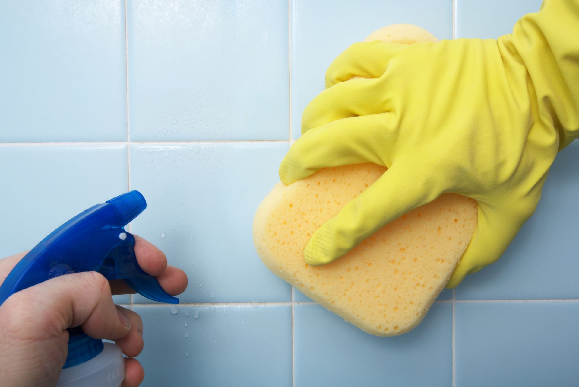 A person wearing yellow gloves is cleaning a tile wall with a sponge.