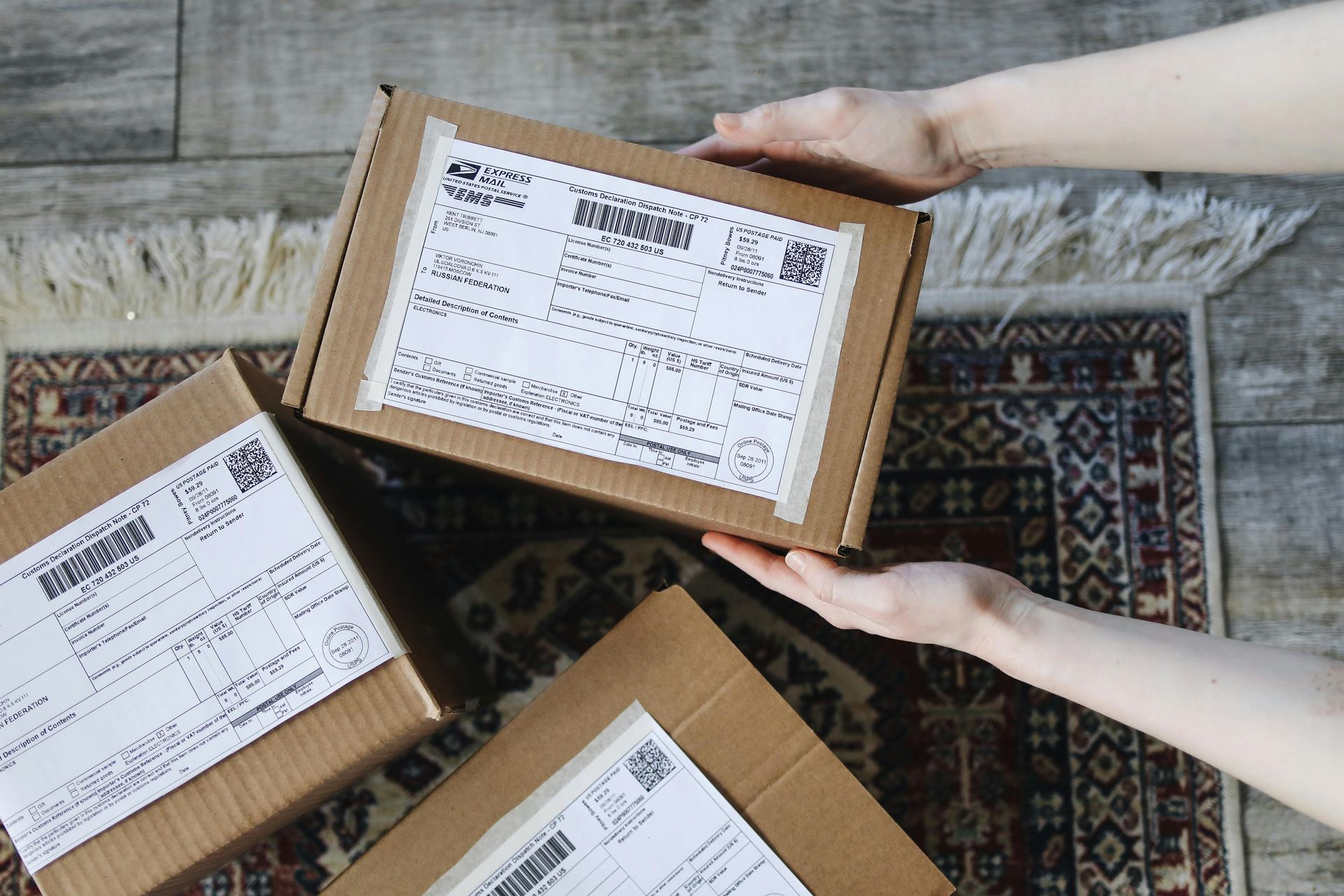 Hands holding a cardboard package with shipping label, surrounded by other packages on a rug.