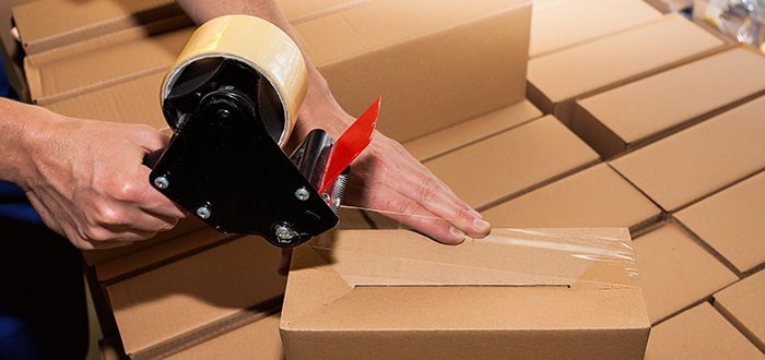 Person using a tape dispenser to seal a cardboard box. Boxes are stacked in a warehouse setting.