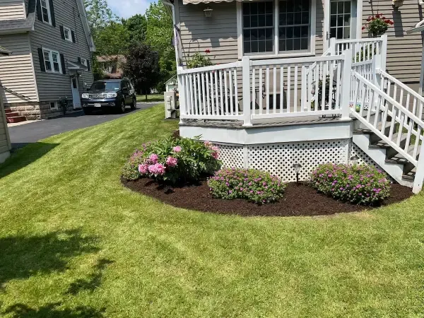 Lawn with mulched flower beds in front of a house with a deck and stairs. A car is parked in the driveway.