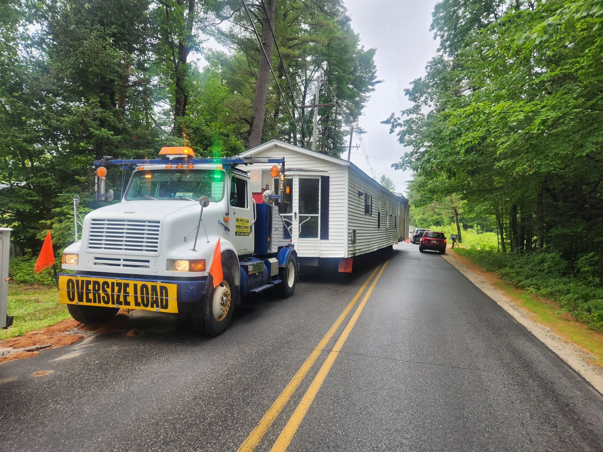 A tow truck transports a mobile home on a narrow road lined with trees. 