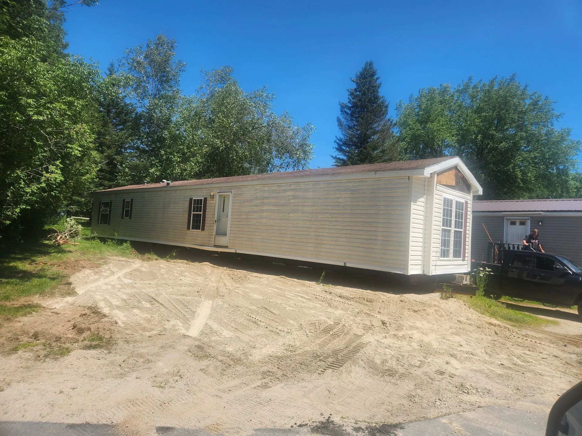 A long, light-colored mobile home sits on a sloped, sandy lot next to green trees and another small building.