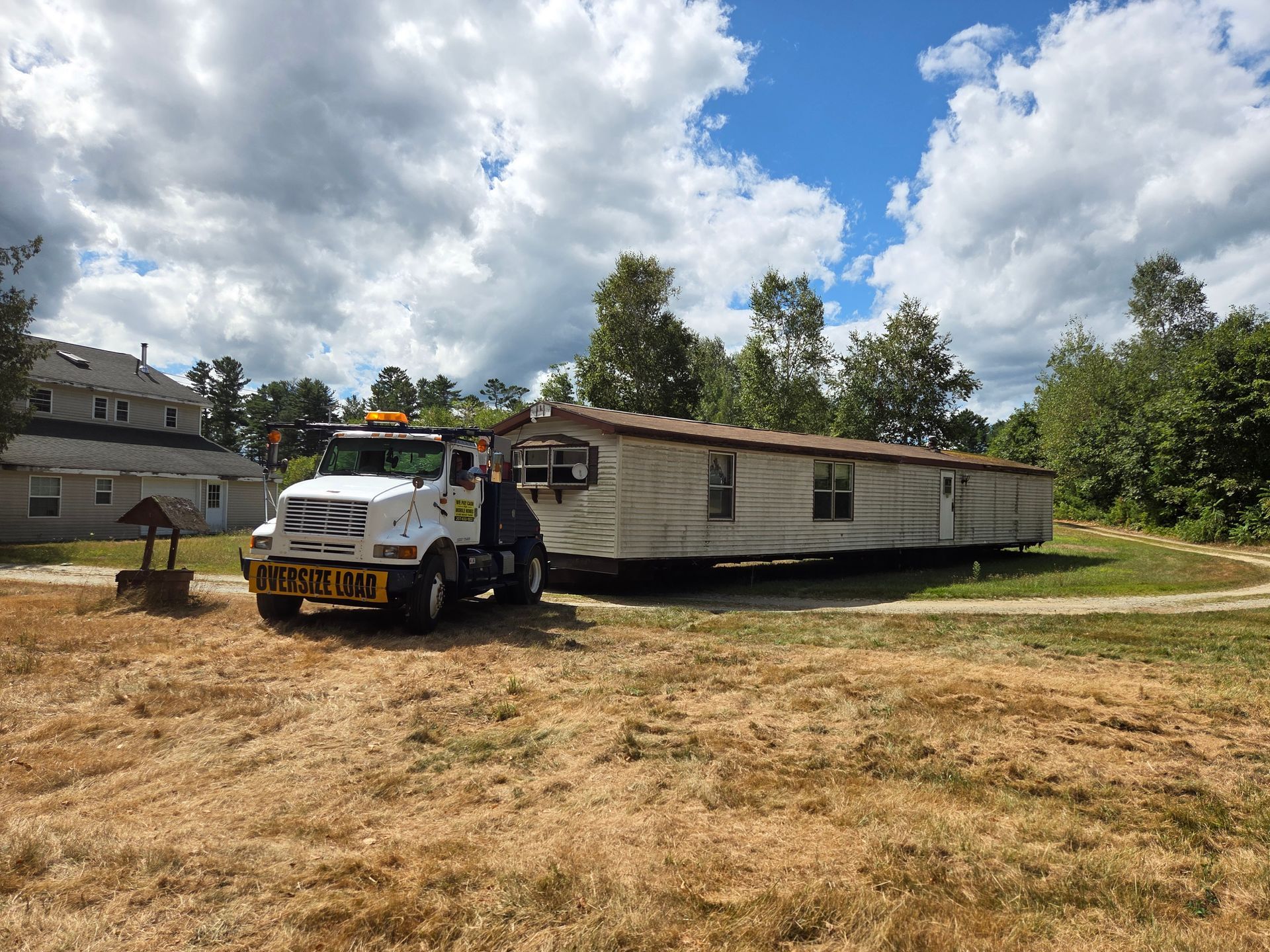 A mobile home being transported by a white truck on a grassy field under a cloudy sky.