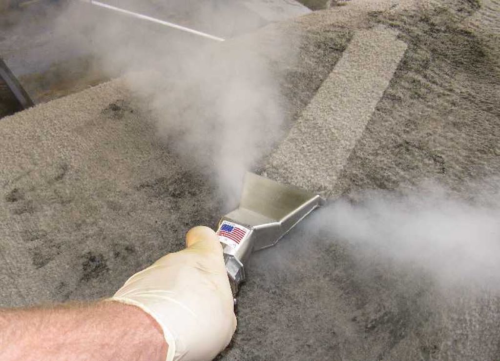 Hand steam-cleaning carpet with a stainless steel nozzle, creating steam, indoors.