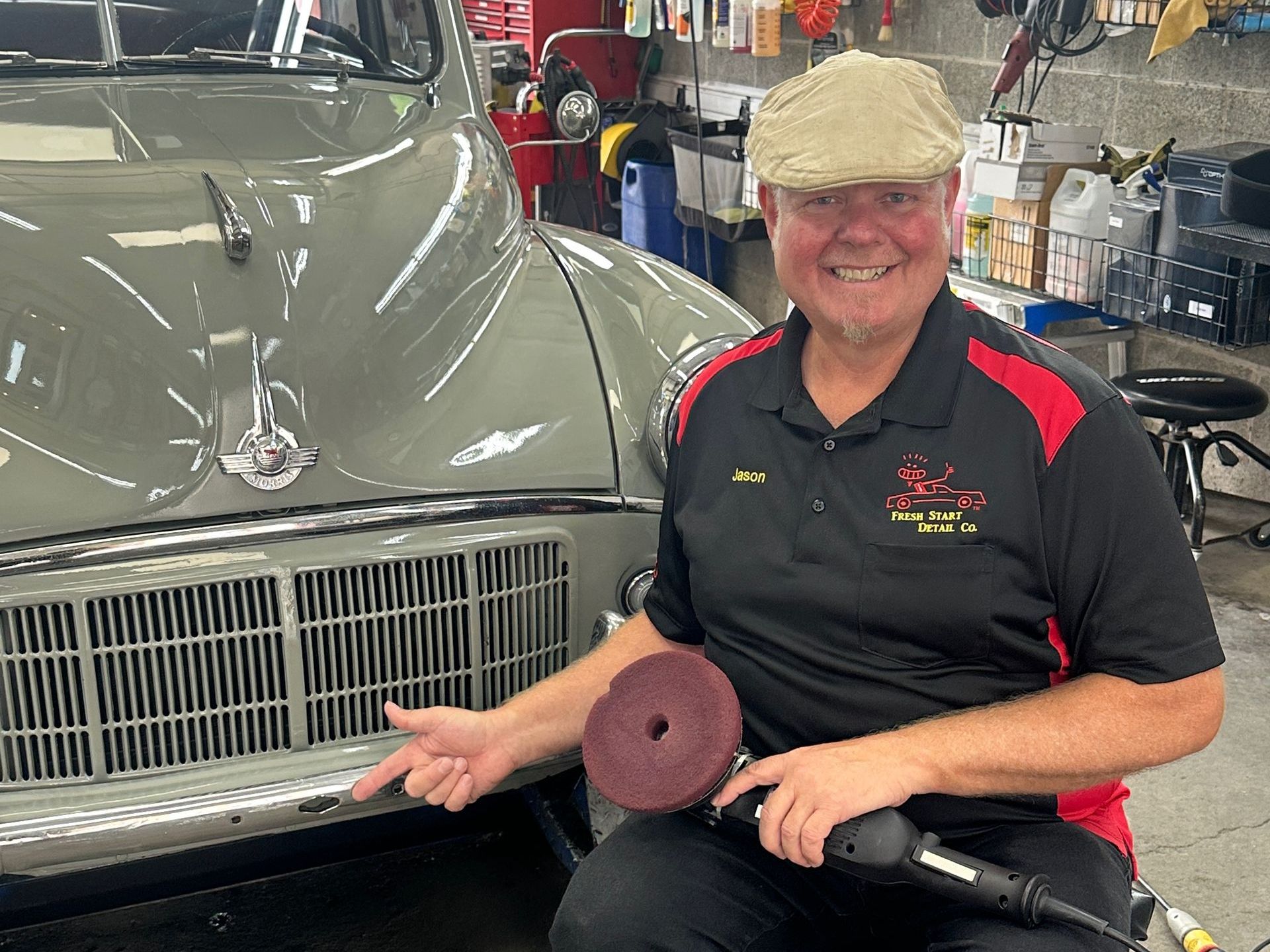 Man in auto shop holds polisher, smiles next to vintage car.