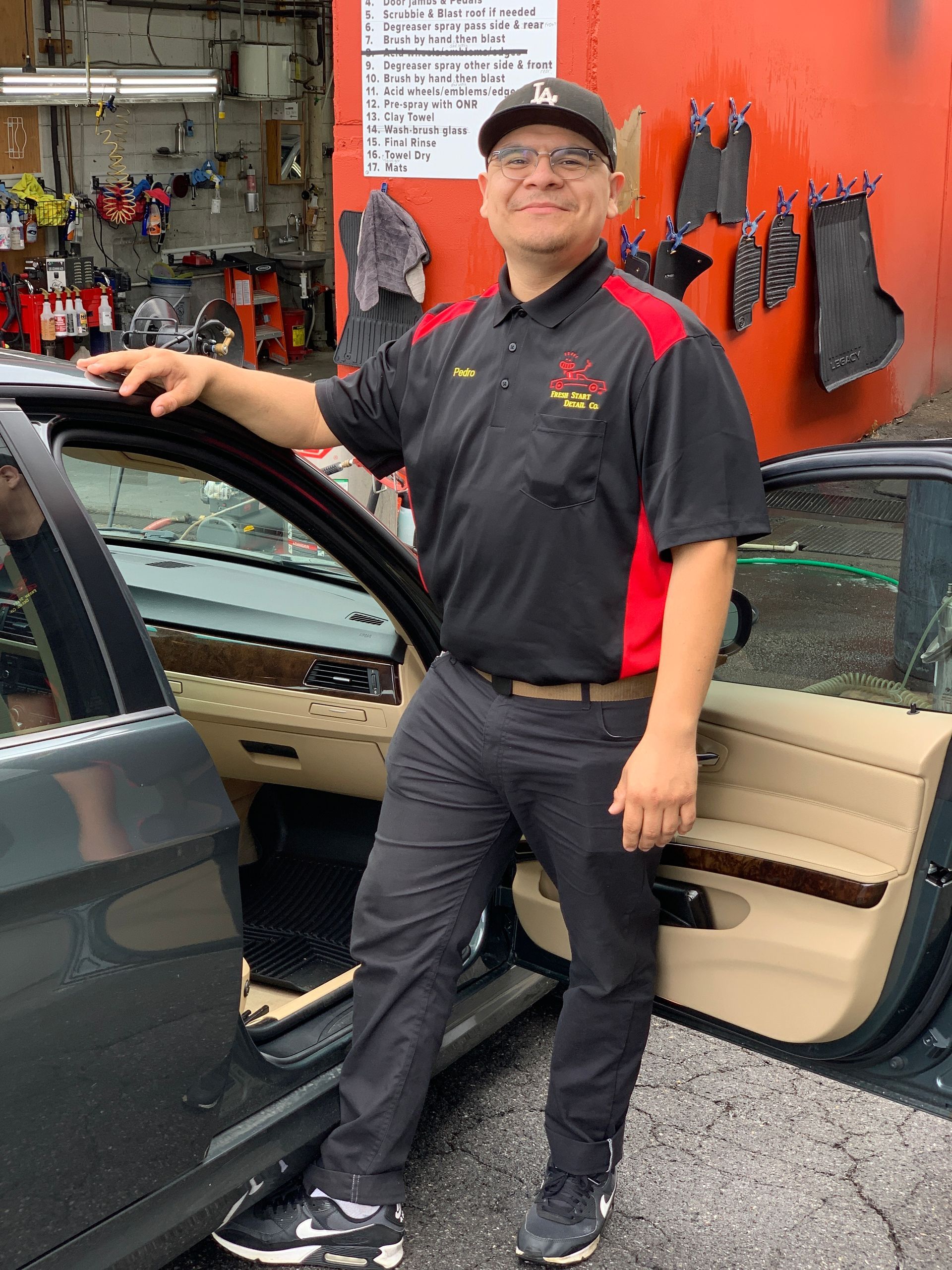 Man in work uniform leans on a car door in an auto shop; he's smiling and wearing a baseball cap.