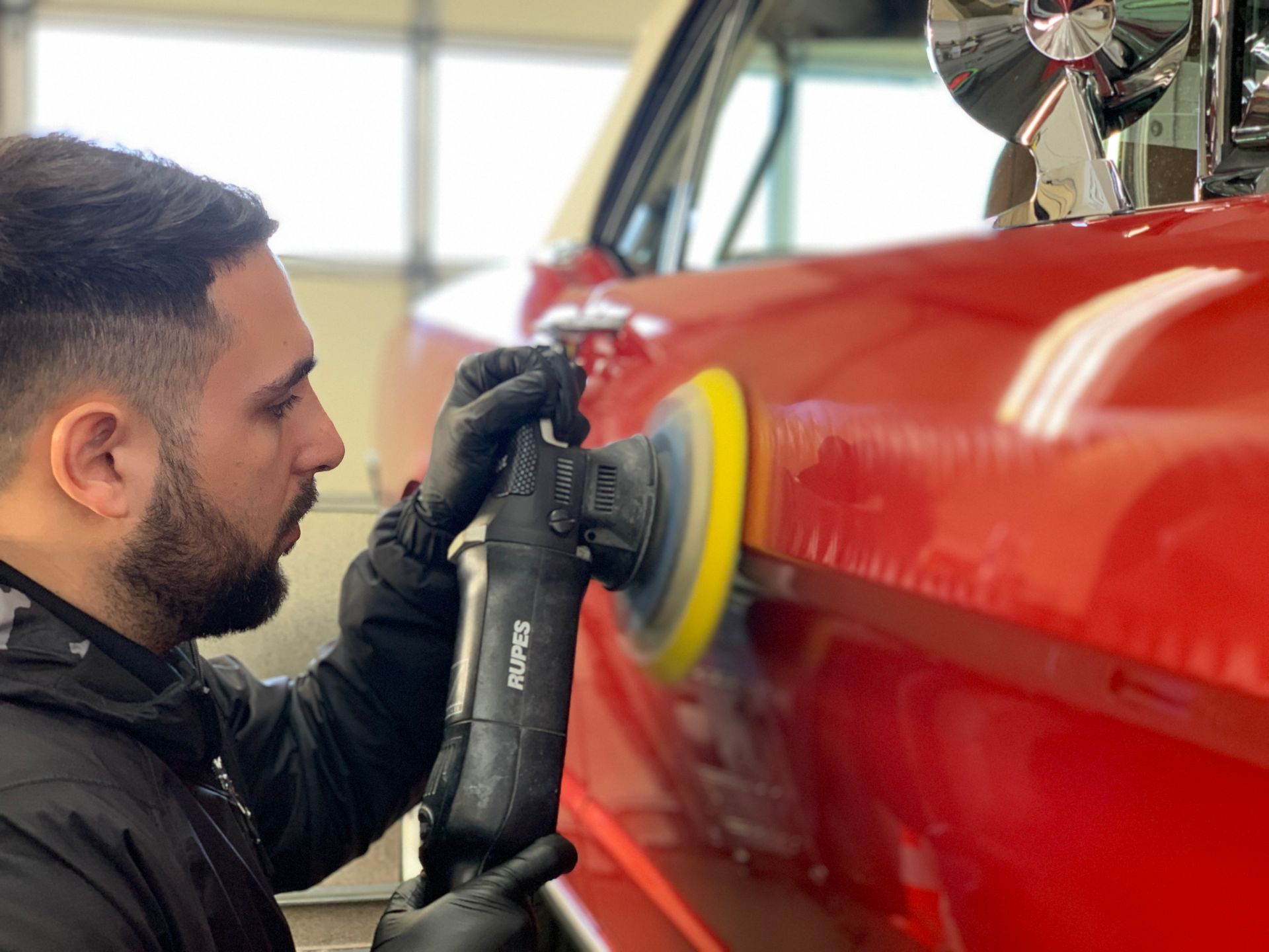 Man in black gloves polishing a red car with a rotary buffer.