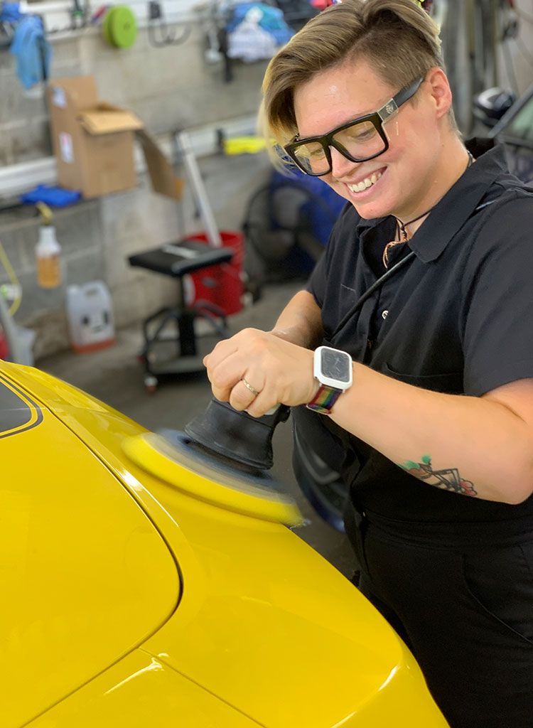 Woman in glasses polishes a yellow car with a buffer in a garage, smiling.