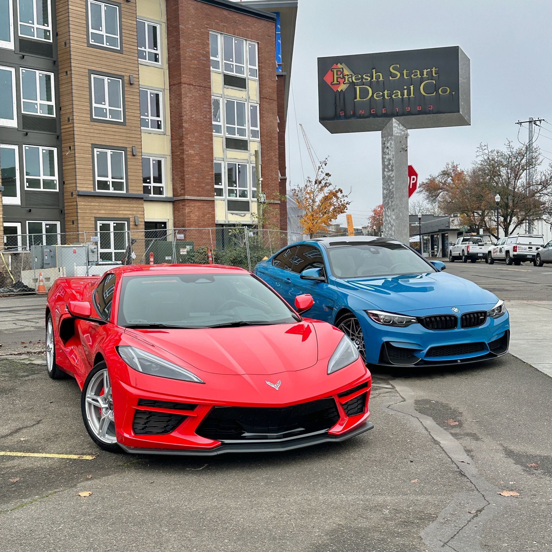 Red Corvette and blue BMW parked in front of a car detailing business. Buildings in background.