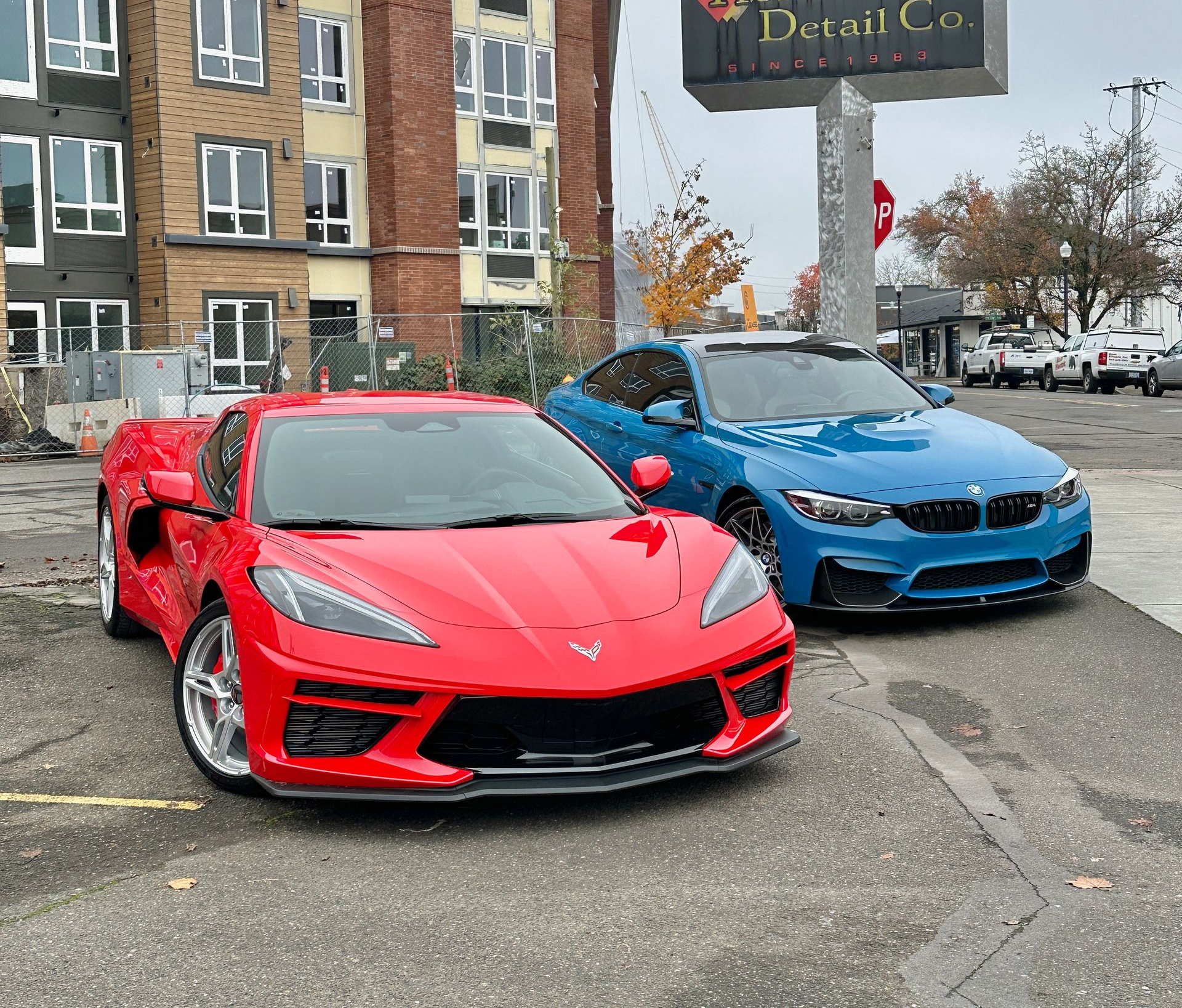 Red Corvette and blue BMW parked in front of a car detailing business. Buildings in background.