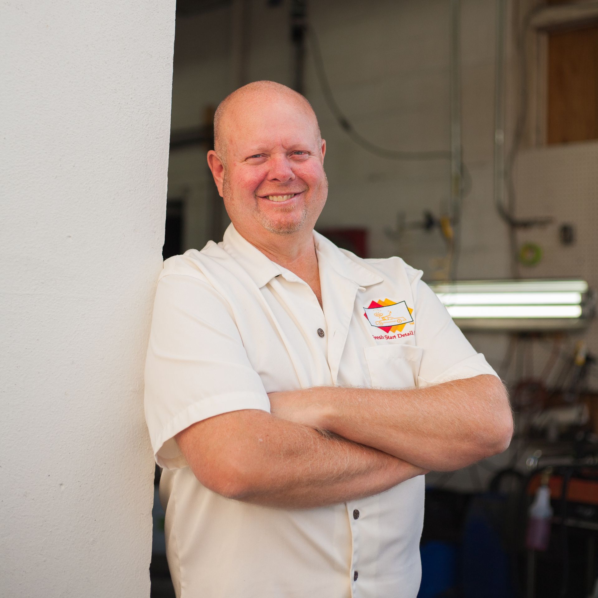 Bald man, arms crossed, smiling, leaning against a white wall in a shop; wearing cream-colored shirt with logo.