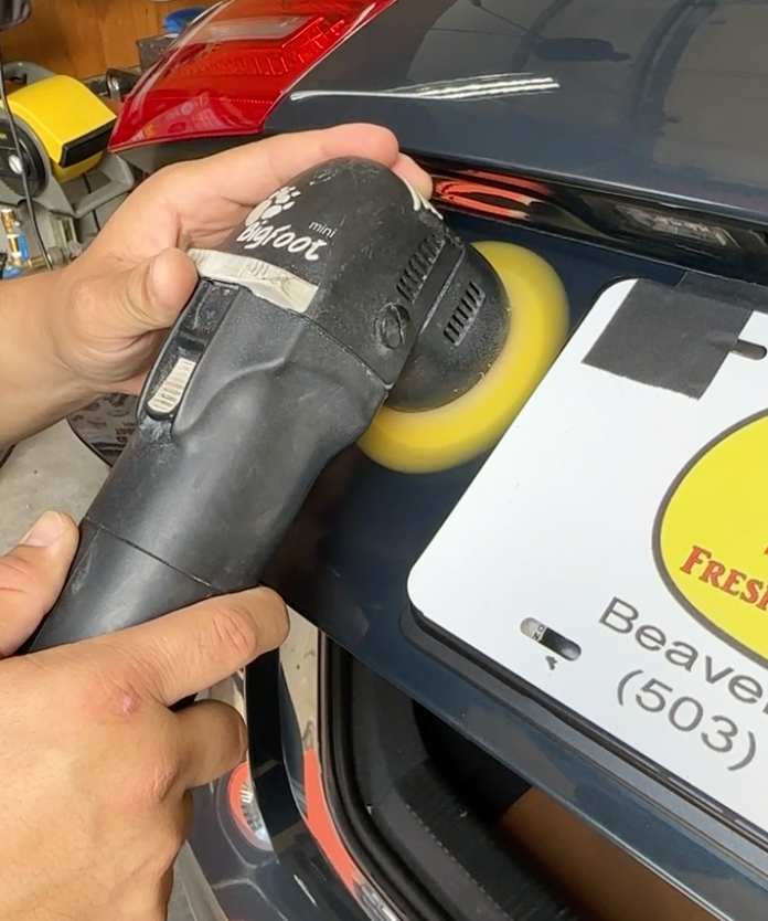 Person using a Bigfoot polisher on a car's rear panel. The license plate is taped for protection.