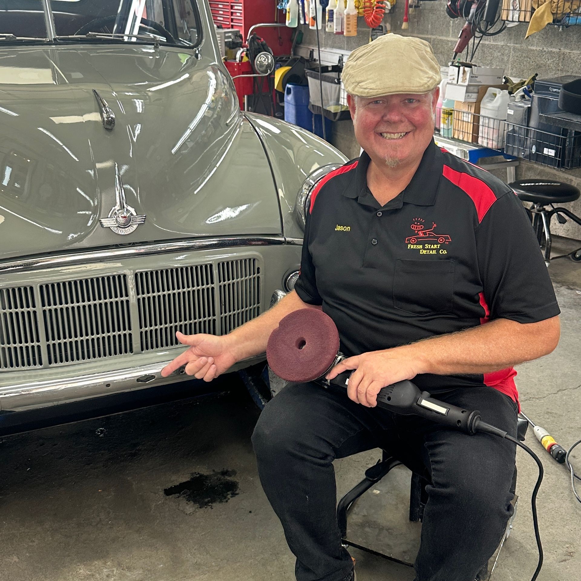Man polishing a vintage gray car's bumper with a buffer. He's wearing a cap and pointing at the car.