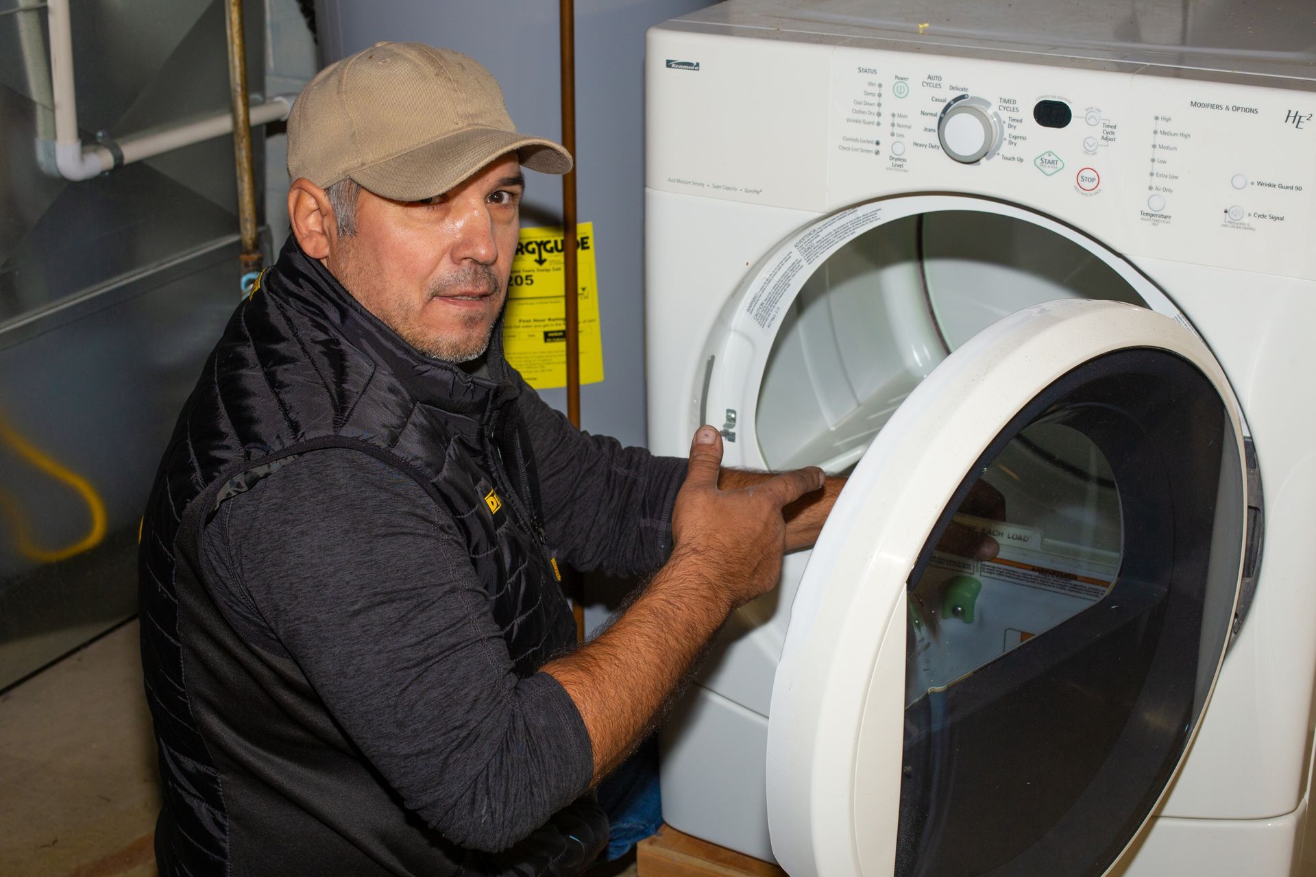Man in cap and vest opens a white dryer door in a utility room.