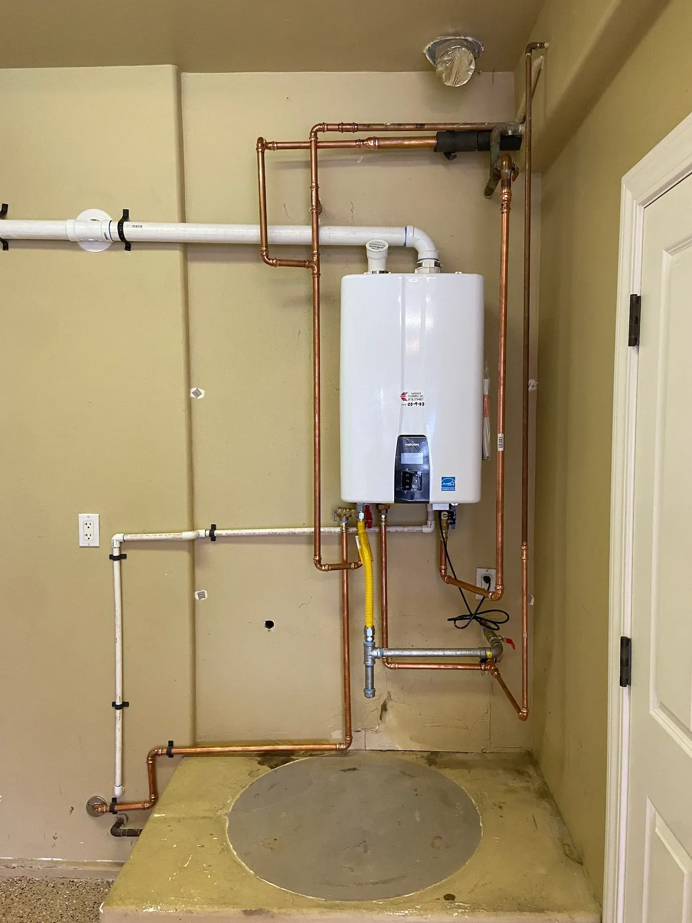 A white water heater is connected to copper pipes in a room.