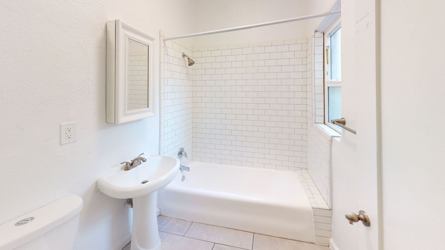 White bathroom with a pedestal sink, tub/shower, small window, and wall-mounted mirror.
