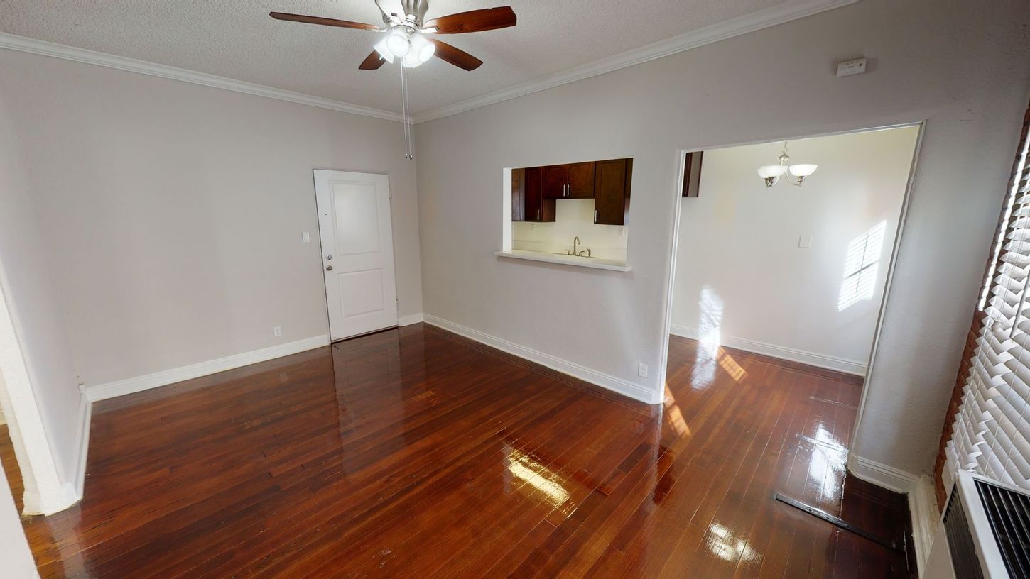 Interior view of a living room with wooden floors, a ceiling fan, and a view into a kitchen area.