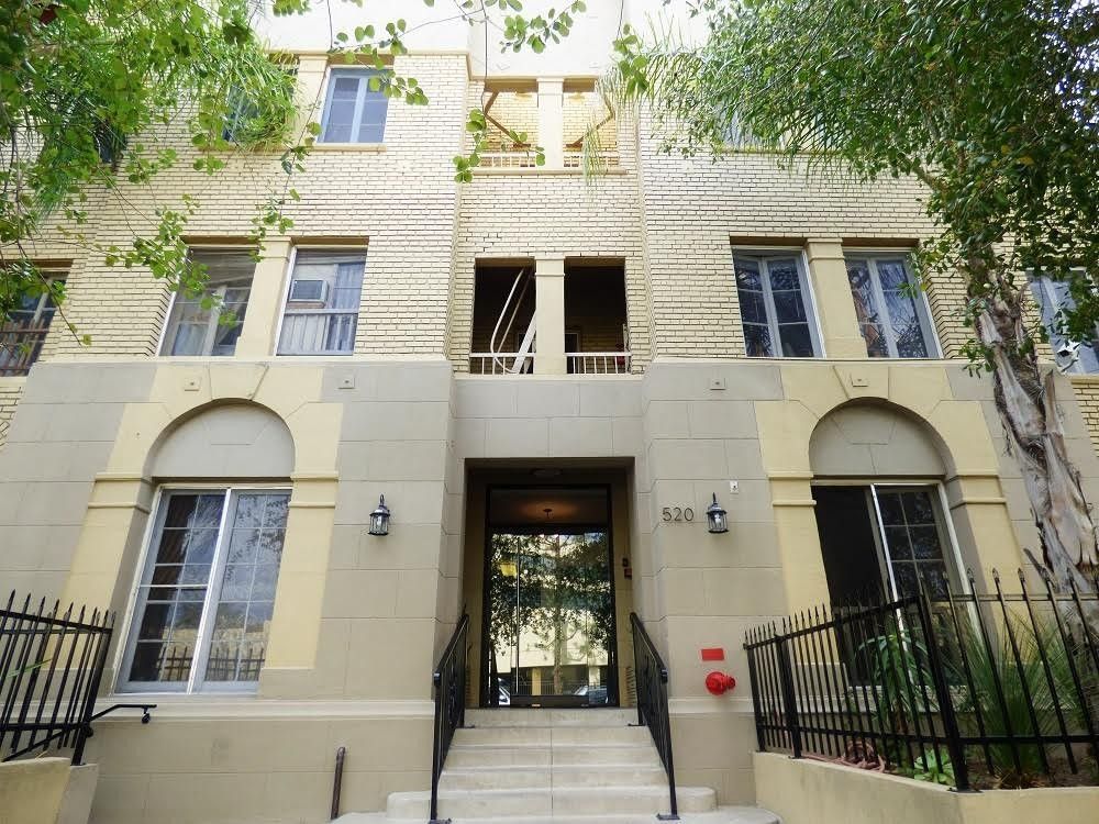 Apartment building exterior, light-colored facade with arched windows and entryway. Black wrought iron railing, trees.