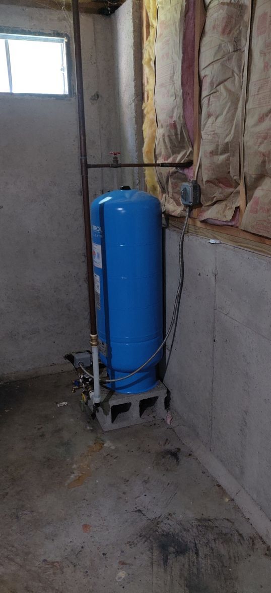 A blue water tank is sitting in a basement next to a window.