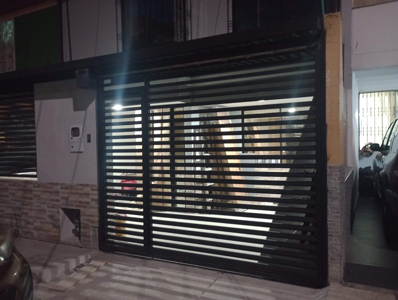 Puerta negra con lamas horizontales que oculta parcialmente el interior de un edificio. Escena nocturna, con luz emanando del interior.