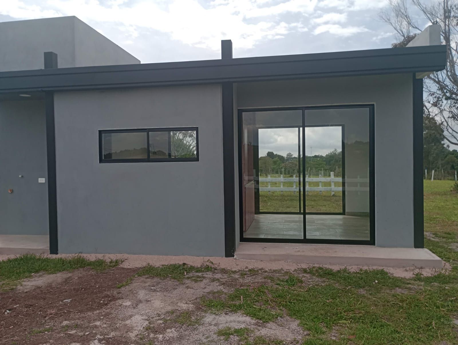 Edificio gris con detalles negros, puertas corredizas de vidrio, ventana pequeña y césped verde afuera.