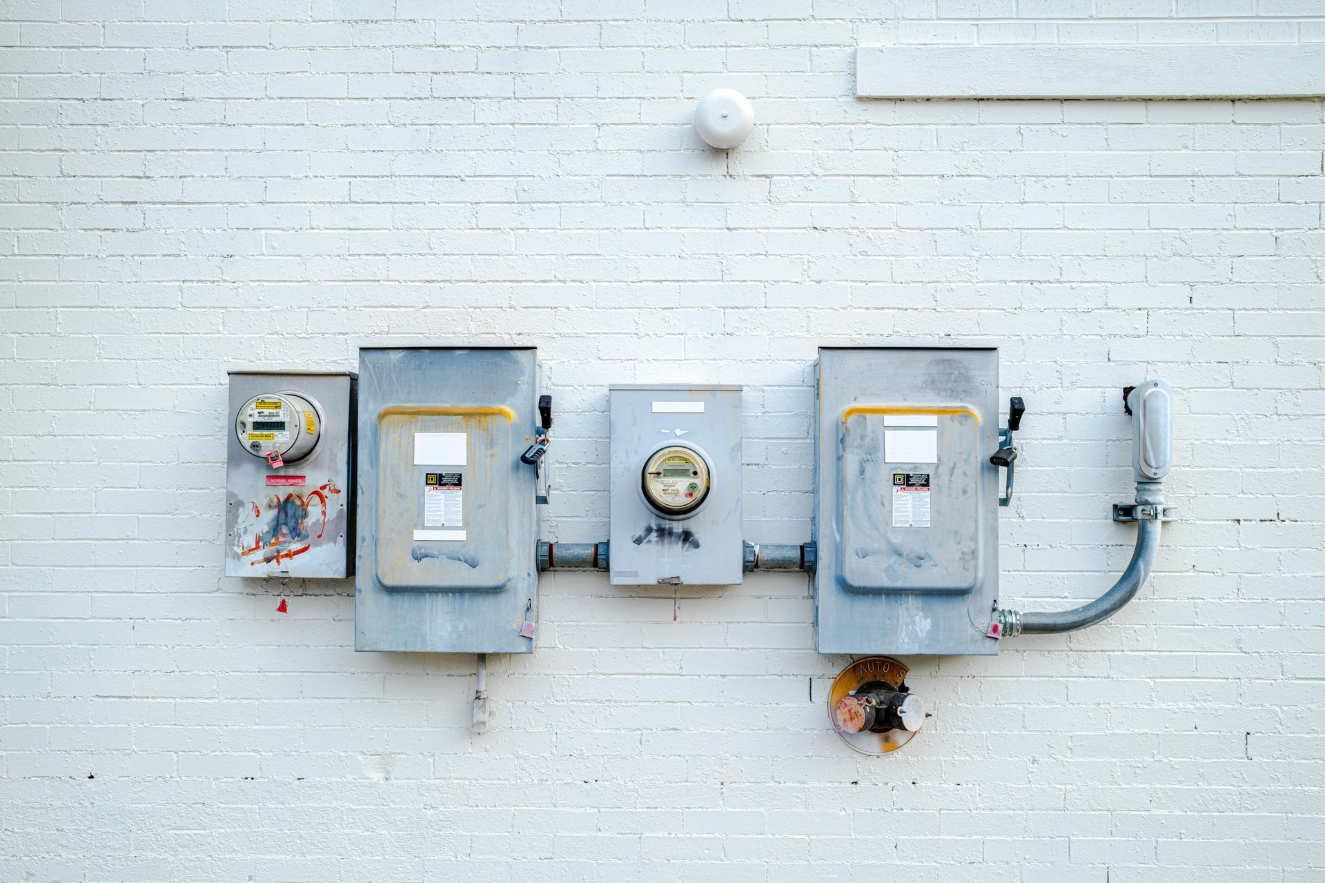Wall with several gray utility meter boxes and electrical panels against white brick, with cables attached